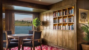 A room with a wooden bookshelf, a dining table with chairs, and a view of a body of water. - Steigenberger Royale Nile Cruise - Every Thursday from Luxor for 07 & 04 Nights - Every Monday From Aswan for 03 Nights (Luxor)