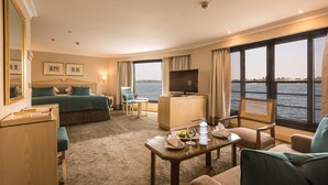A hotel room with a bed, a desk, a chair, a TV, and a view of the water. - Steigenberger Royale Nile Cruise - Every Thursday from Luxor for 07 & 04 Nights - Every Monday From Aswan for 03 Nights (Luxor)