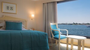 A hotel room with a bed, a chair, a small table, a nightstand, a lamp, and a view of the water and boats. - Steigenberger Royale Nile Cruise - Every Thursday from Luxor for 07 & 04 Nights - Every Monday From Aswan for 03 Nights (Luxor)