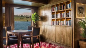 Quarto com uma grande janela com vista para um lago, uma estante de madeira repleta de livros, uma mesa de jantar com cadeiras e um tapete vermelho estampado.