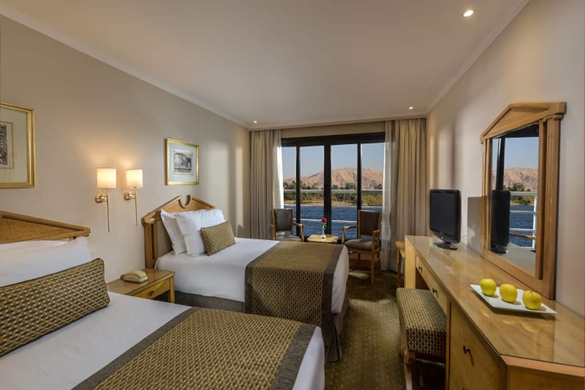 A hotel room with two beds, a desk with a television, and a view of a body of water and mountains.