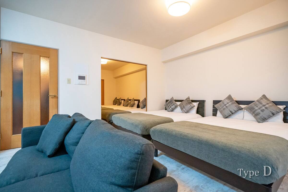 Habitación de hotel con dos camas individuales, un sofá, una puerta de madera y una lámpara en el techo.
