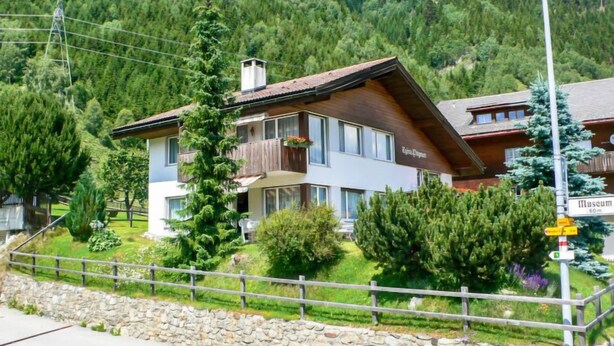 A two-story chalet with a wooden facade, balconies with flower boxes, and a stone wall in the foreground.