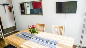 A dining area with a wooden table, chairs, a vase with a flower, a checkered placemat, a mirror, and a TV on the wall.