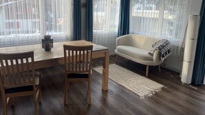 A living room with a wooden table, two chairs, a sofa, and a large window with sheer curtains.