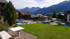 A parking area with several cars, a grassy lawn, and buildings with mountain views in the background.