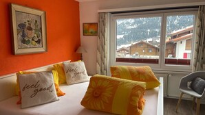A hotel room with a bed, pillows with 'feel good' text, a painting, a lamp, and a window with a view of houses and mountains.