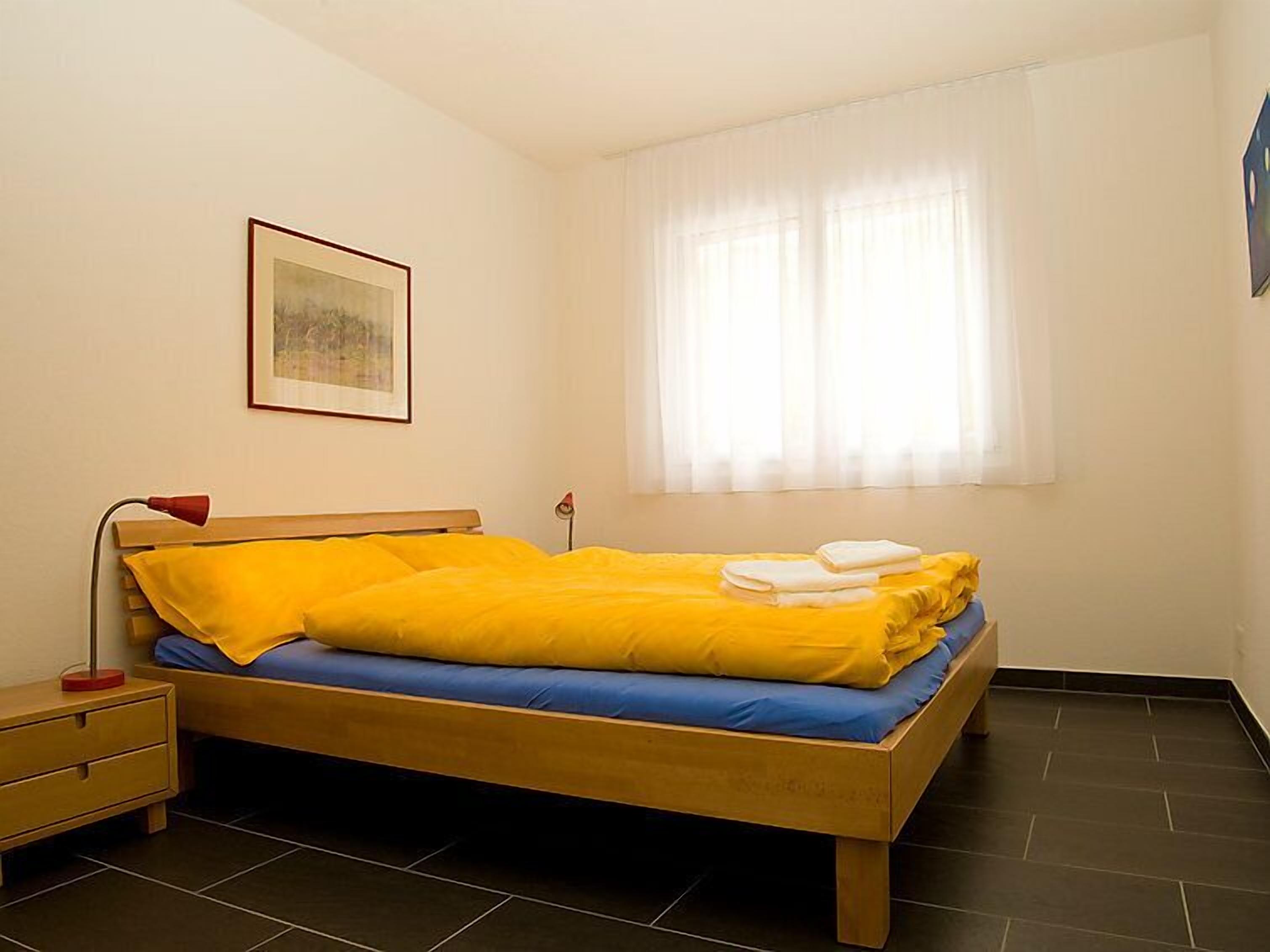 Un lit simple avec une literie jaune et bleue, une table de chevet, une lampe et un tableau encadré au mur.