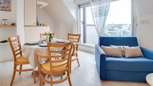 A modern living room with a dining area, featuring a blue sofa, wooden chairs, a dining table set for four, and a large window with curtains. - Bleue Océane by Interhome (Carnac)