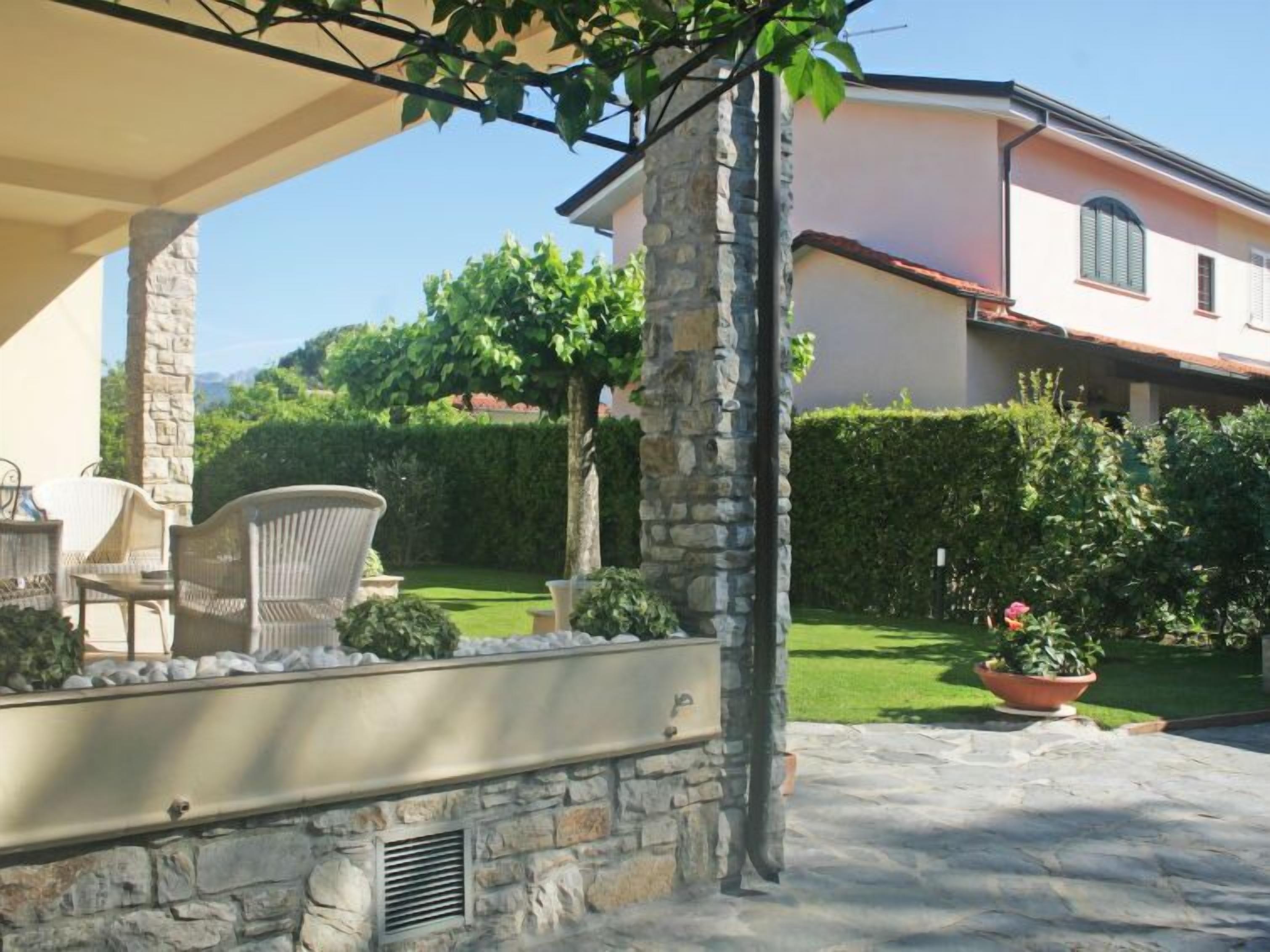 Un patio de piedra con mobiliario exterior, un muro de piedra y un jardín bien cuidado con seto y un árbol.