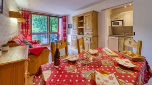 Une pièce chaleureuse comprenant un lit, une table à manger dressée pour le repas, un coin cuisine avec des placards en bois et une grande fenêtre donnant sur la forêt.