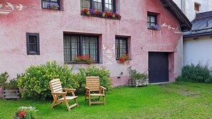 Un bâtiment rose avec un jardin, deux chaises en bois et des jardinières aux fenêtres.