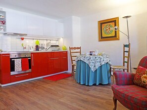 A compact kitchen with red cabinets, a small table with a blue tablecloth, and a wooden chair. - Chesa Rossa by Interhome (Bever)
