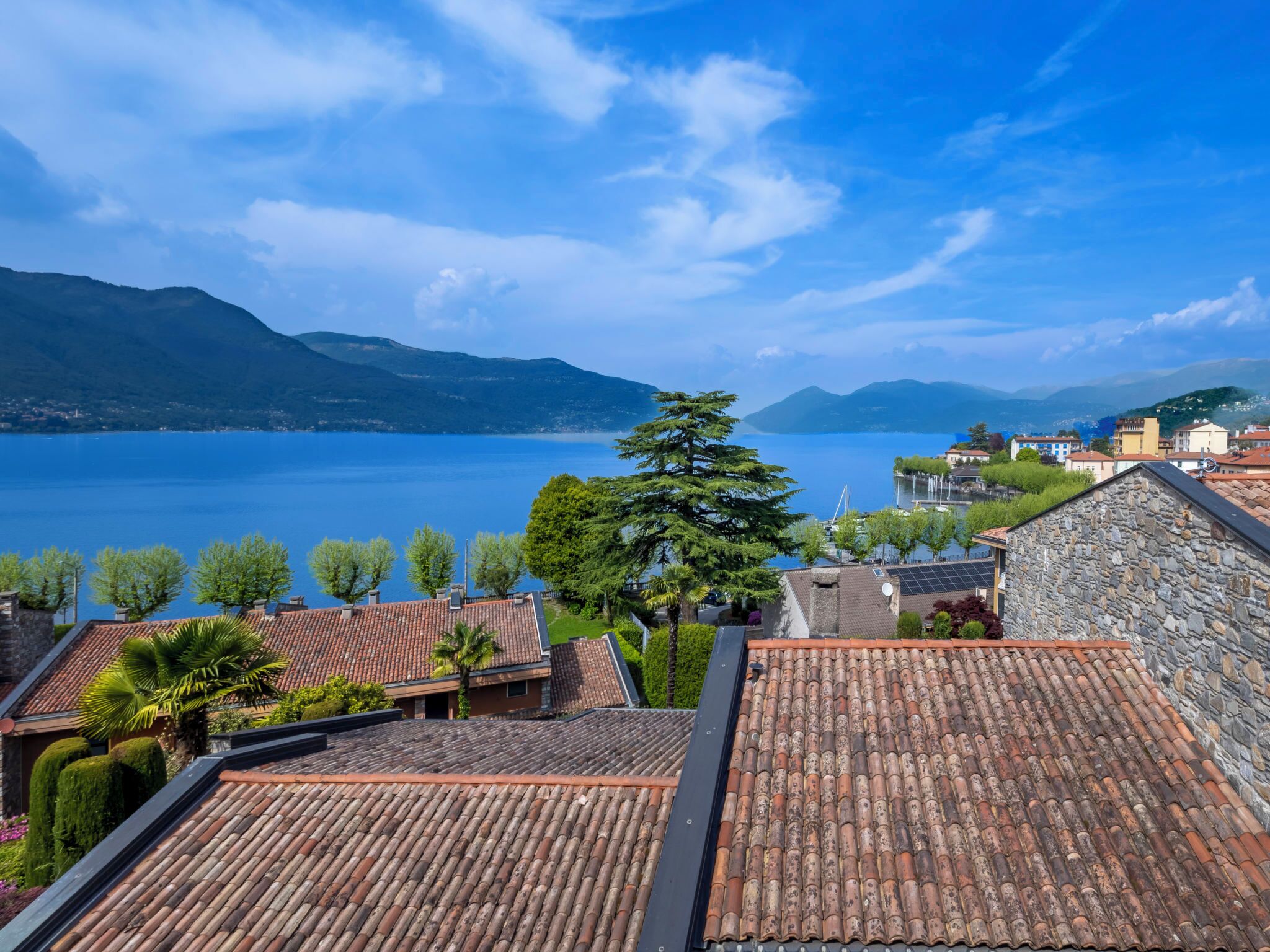 A coastal town with terracotta-roofed buildings, a clear blue lake, and mountains in the background.