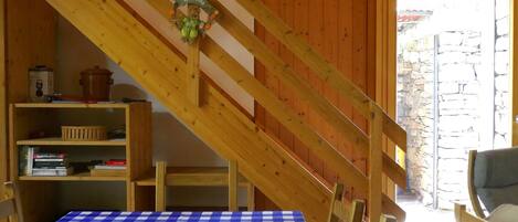 A room with a checkered table, wooden staircase, and a view of a stone wall outside.