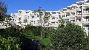 A modern multi-story hotel with balconies, surrounded by lush greenery and a clear blue sky.