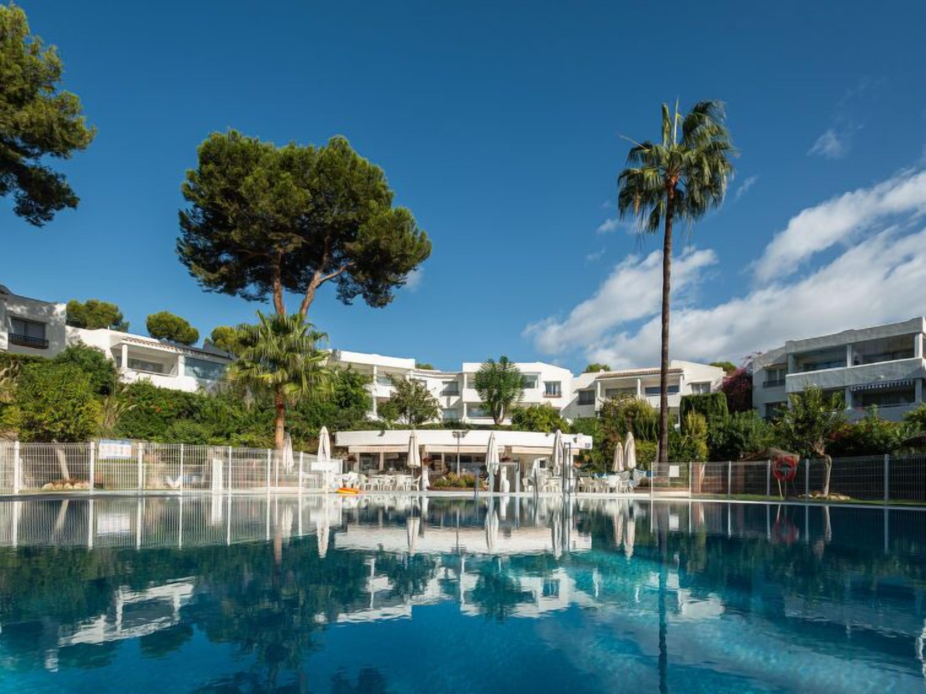A swimming pool with a clear blue sky, surrounded by white buildings and greenery.