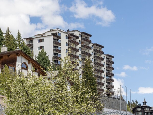 A multi-story residential building with balconies, surrounded by trees and a clear sky.