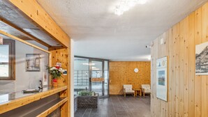 A reception area with a wooden counter, a reception desk, a seating area with two chairs, a clock, and a poster on the wall. - Guardaval Apt 505 by Interhome (Davos)