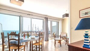 Un coin repas avec vue sur la mer, des chaises en bois, une table dressée pour deux personnes et une lampe à rayures bleues et blanches.