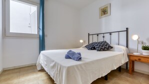 A neatly made bed with a white bedspread, two pillows, and a blue towel. There is a bedside table with a lamp and a small potted plant. A window with a blue curtain is visible, and a framed picture hangs on the wall. - Platja de Roses by Interhome (Roses)