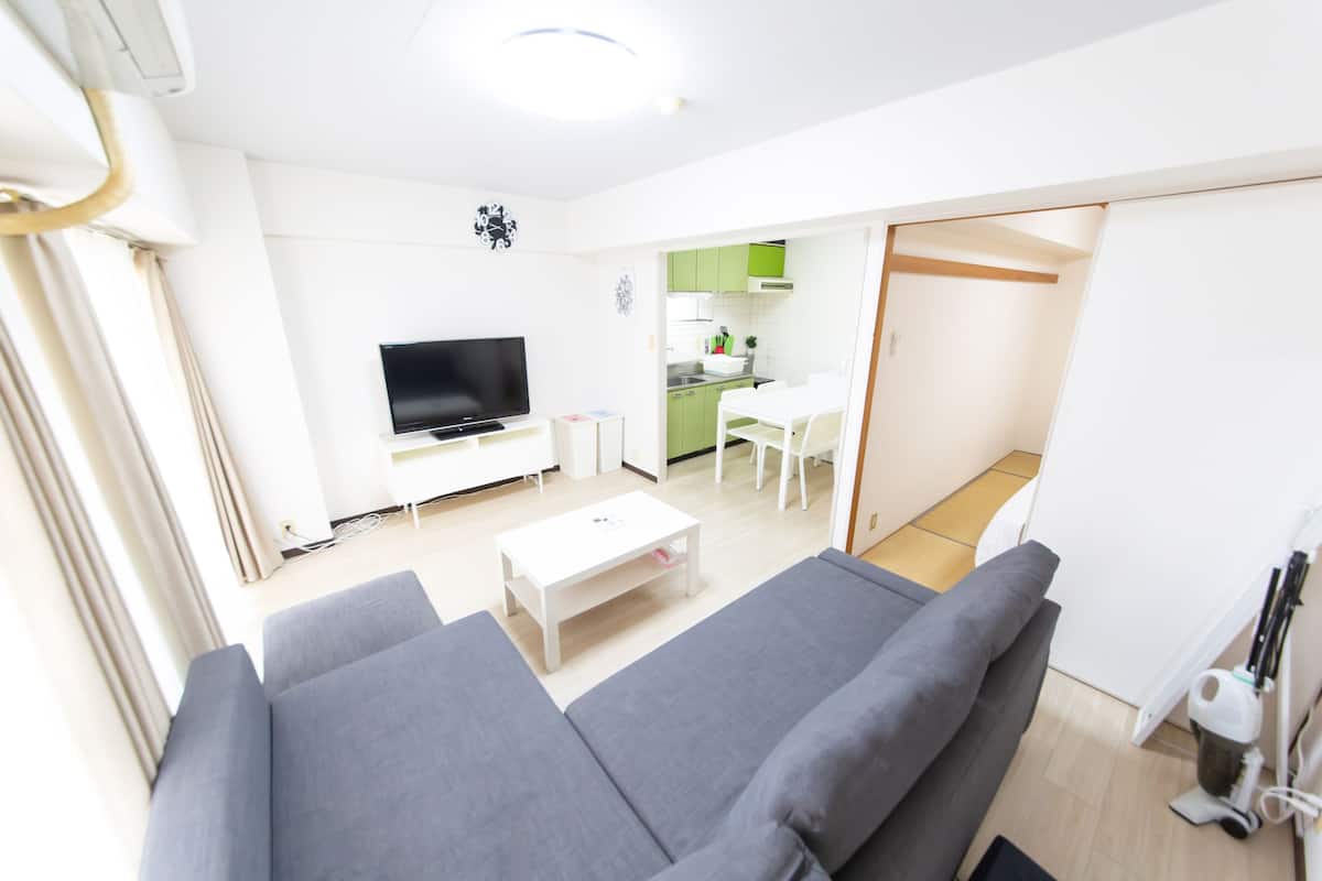 A modern living room with a grey sofa, a white coffee table, and a flat-screen TV. There is a kitchen area with green cabinets and a white countertop visible through an open door.