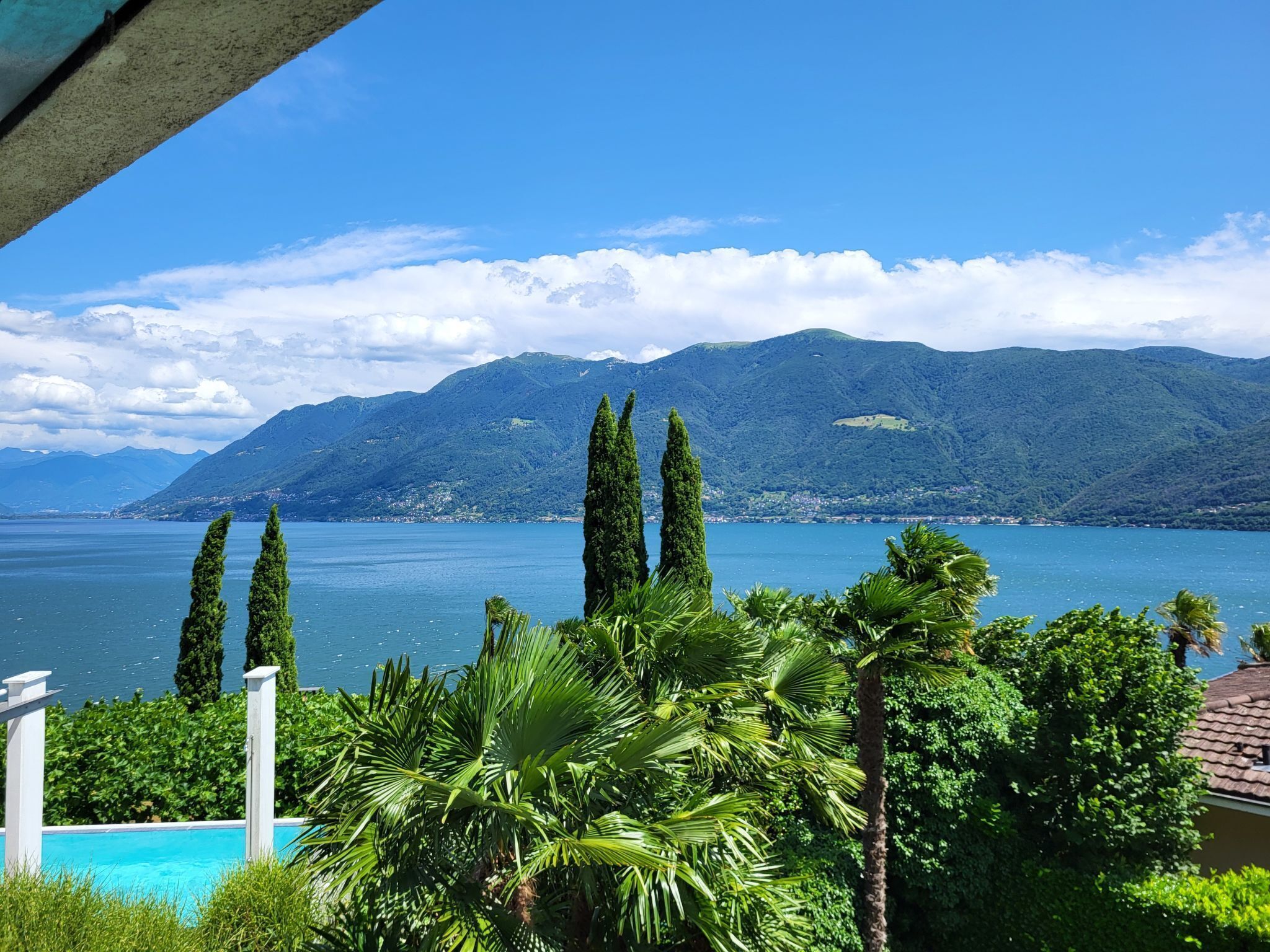 Une vue sur un lac avec des montagnes en arrière-plan, un espace piscine bordé de palmiers et un ciel dégagé.
