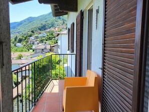 A balcony with a view of a hillside village, a closed shutter, and a yellow chair.