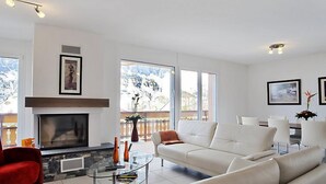 A modern living room with a fireplace, white sofas, a glass coffee table, and large windows offering a view of the outdoors. - Apartment A31 by Interhome (Leukerbad)