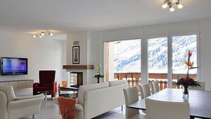 A modern living room with a sofa, TV, fireplace, dining table, and large windows with a snowy view. - Apartment A31 by Interhome (Leukerbad)