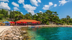 A coastal area with clear turquoise water, a rocky shoreline, and a red-roofed building with outdoor seating. - Apartment Delfin by Interhome (Karlobag)