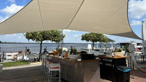 An outdoor bar area with a wooden deck, white bar stools, and a large canopy providing shade. - Busch en Dam by Interhome (Uitgeest)