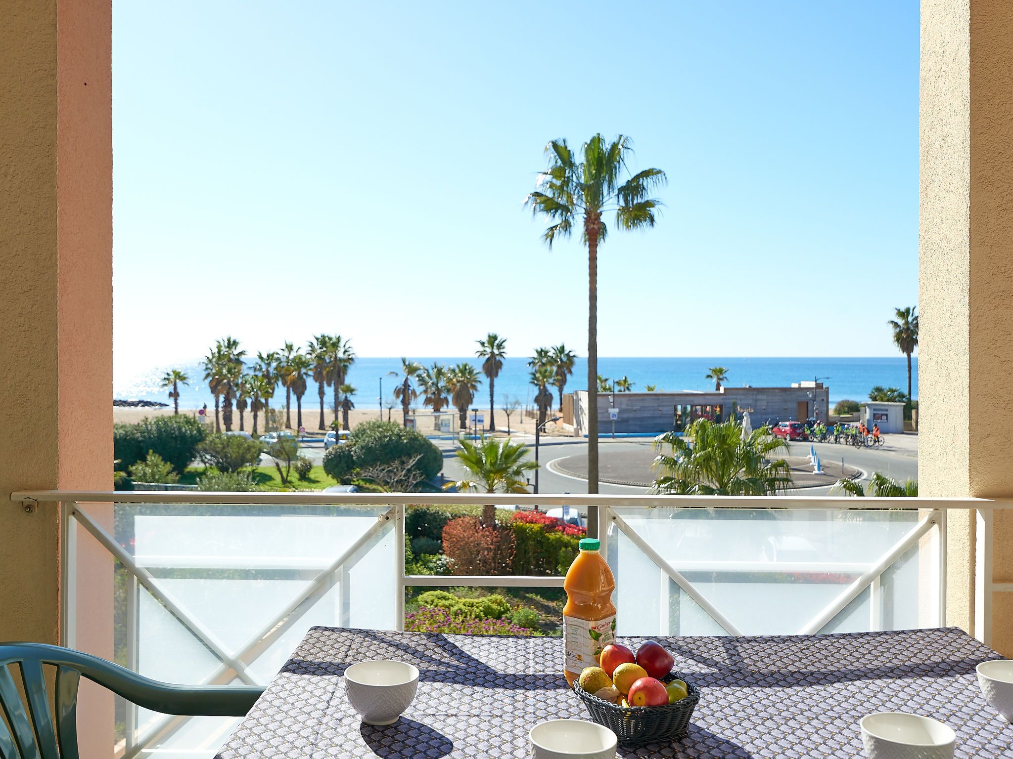 A balcony with a table set for two, overlooking a beach with palm trees and a clear blue sky.