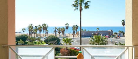 A balcony with a table set for two, overlooking a beach with palm trees and a clear blue sky.