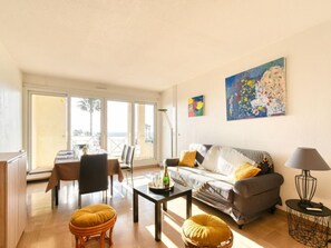 A modern living room with a sofa, a dining table, and chairs. There is a large window providing a view outside.