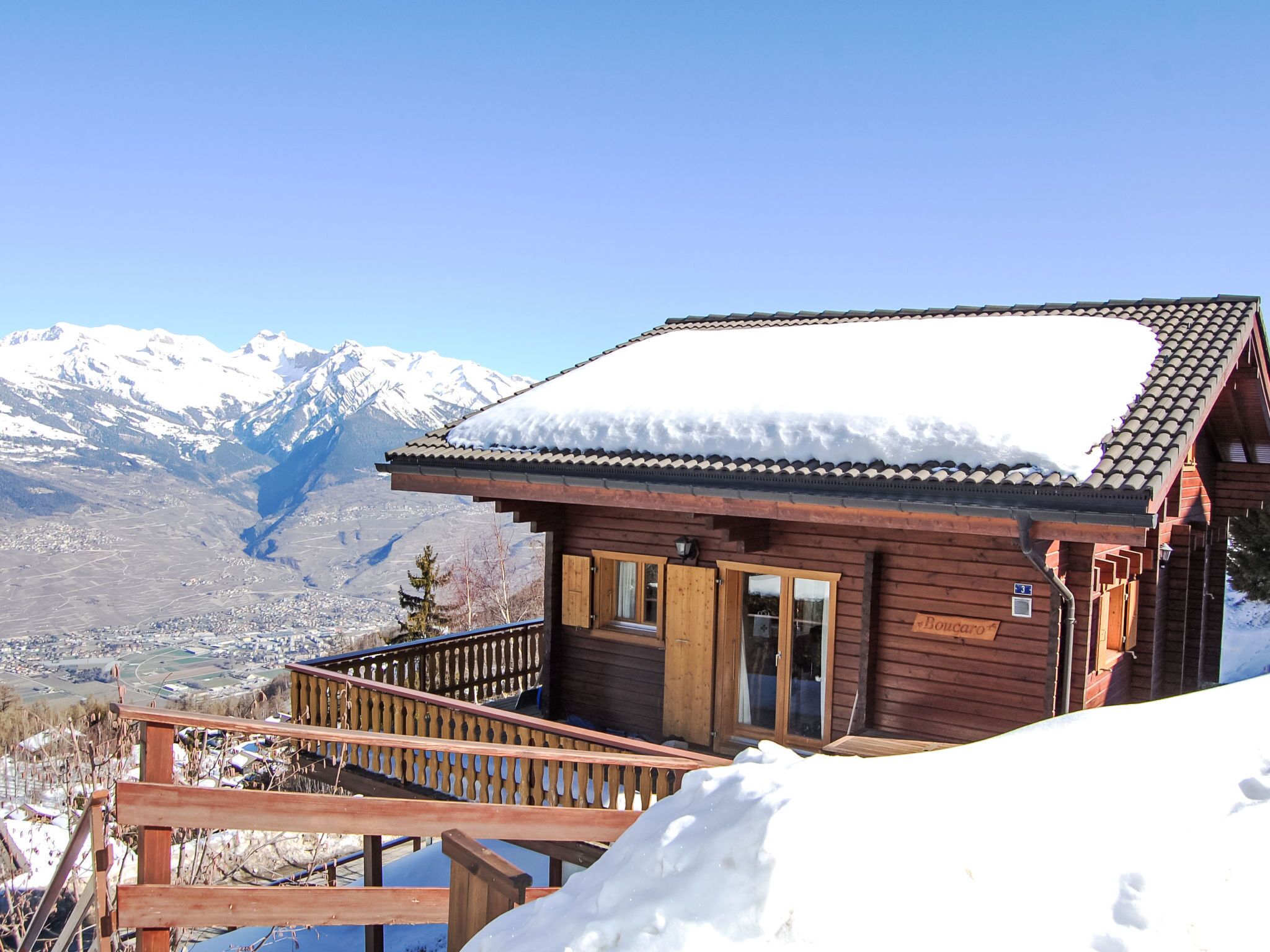 Eine Holzhütte mit schneebedecktem Dach, einem Holzbalkon und Blick auf verschneite Berge.