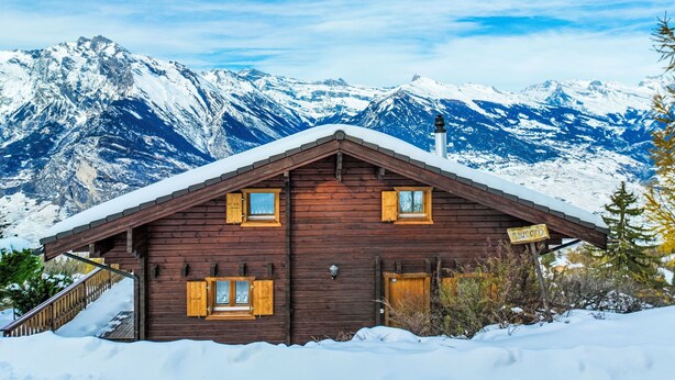 Uma cabana de madeira com telhado inclinado, venezianas de madeira e chaminé, rodeada por montanhas cobertas de neve.