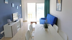 Un salon moderne avec un téléviseur, des meubles blancs, une table à manger et un balcon agrémenté d’un canapé bleu.