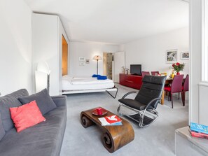 A modern hotel room with a bed, a sofa, a dining table with chairs, a TV, and a desk.