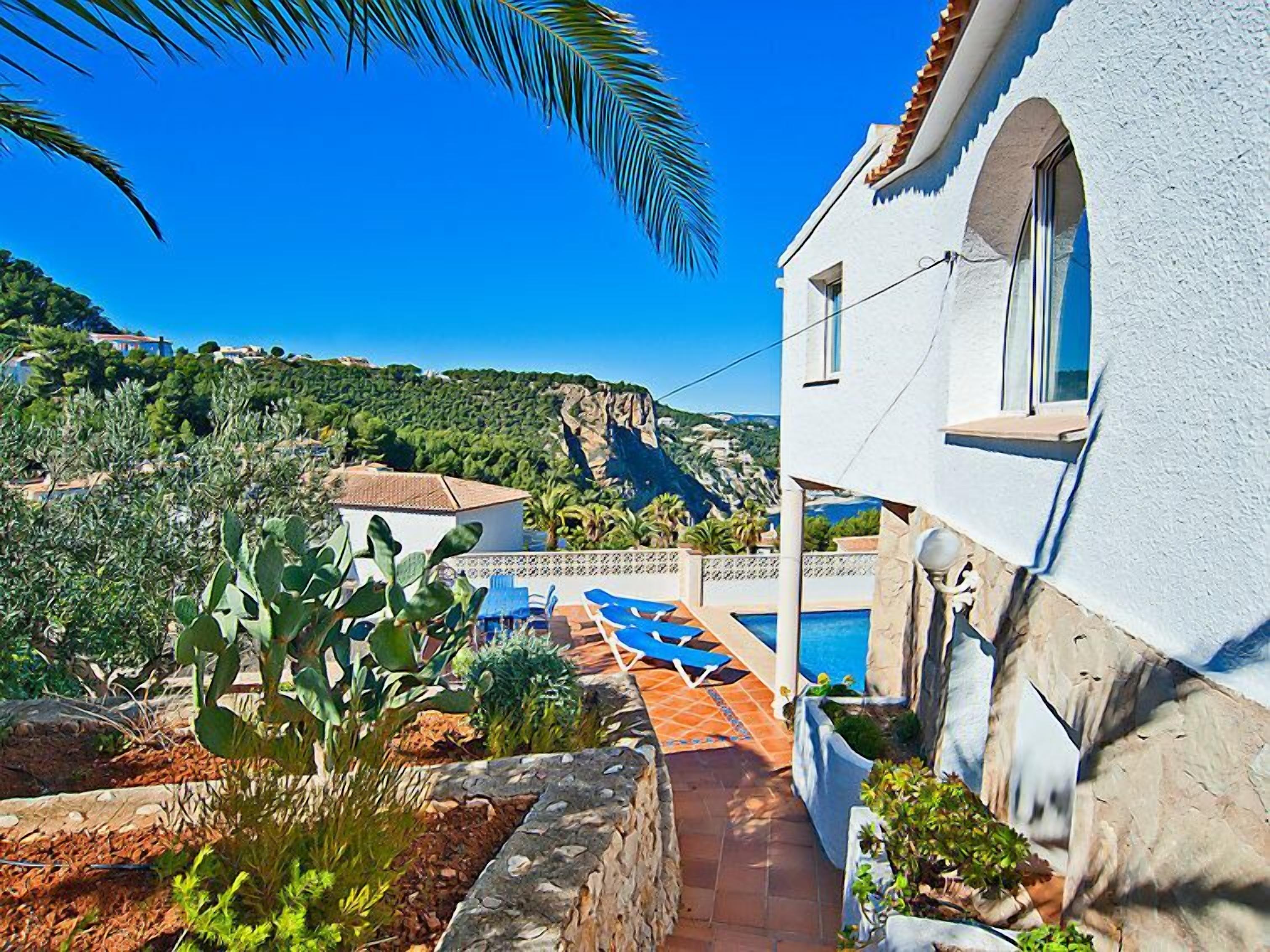 A white villa with a pool, surrounded by greenery and a view of a mountainous landscape.