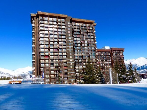 A multi-story hotel with balconies, located near a body of water and mountains.