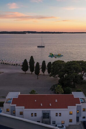A building with a red-tiled roof, a swimming pool, and a view of a calm body of water with a sailboat and inflatable raft.