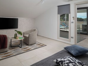 Un salon moderne doté d’un téléviseur à écran plat, d’un fauteuil confortable, d’une petite table avec une plante et offrant une vue sur un balcon aménagé avec du mobilier d’extérieur.