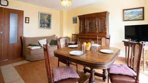 Een eetgedeelte met een houten tafel, stoelen en een kroonluchter. Achterin staat een bank en een kast.