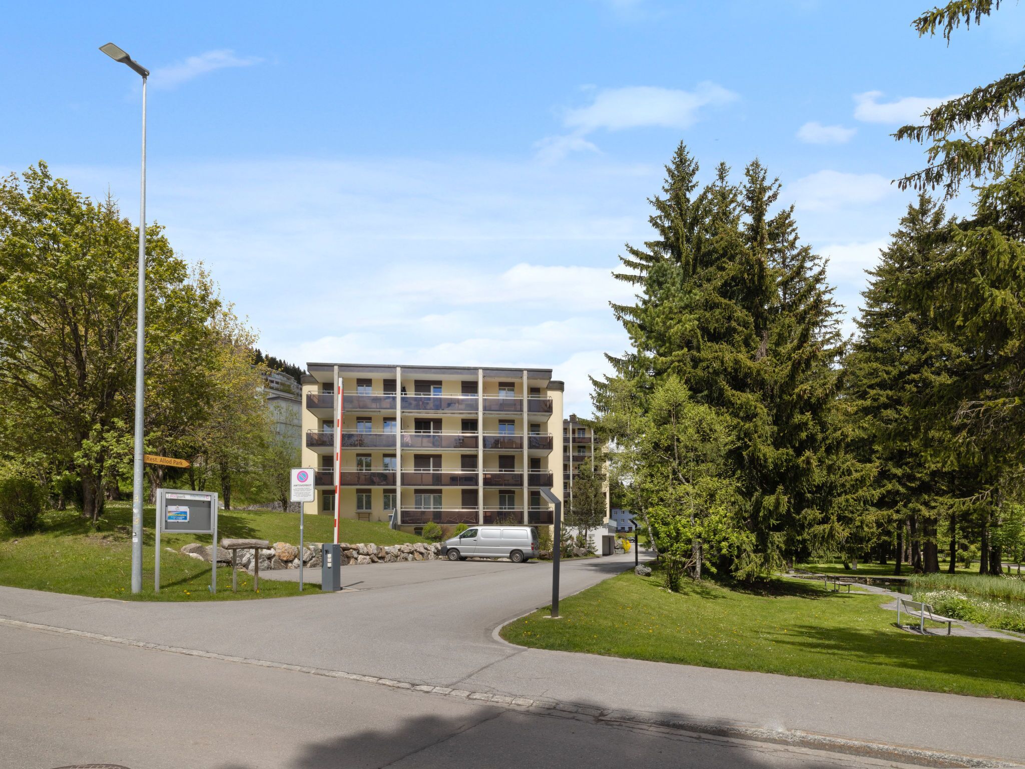 A multi-story building with balconies, a parking area, and a pathway leading to a park.
