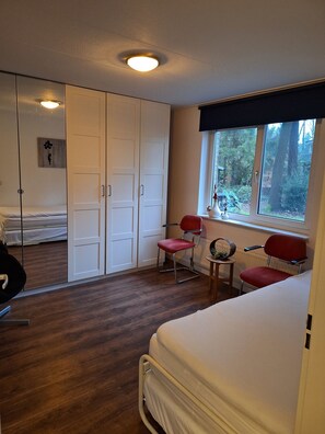 Une chambre à coucher comprenant un lit, une armoire, une chaise, une petite table et une fenêtre donnant sur des arbres.