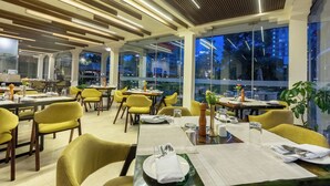 A modern dining area with yellow chairs, tables set with white tablecloths, and large windows offering a view of the cityscape. - The Malla Hotel (Kathmandu)