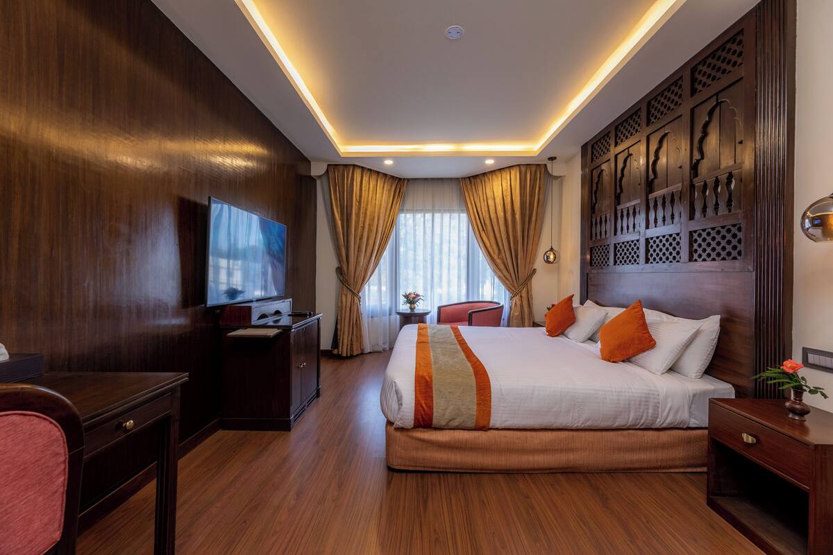 A hotel room with a large bed, wooden paneling, a desk with a TV, and a small table with a vase.