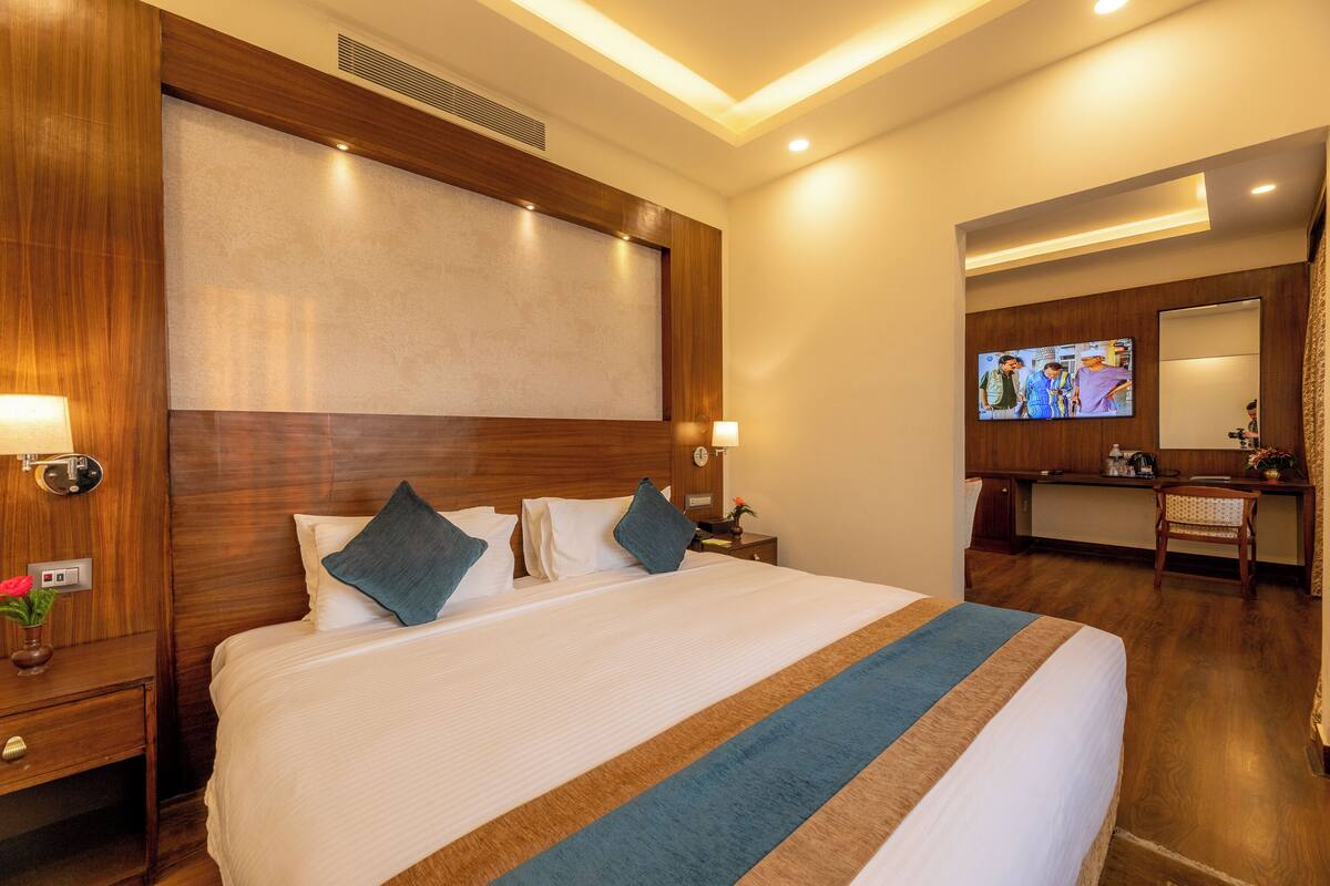 A hotel room with a large bed, wooden headboard, bedside tables, a TV, a desk, and a mirror.