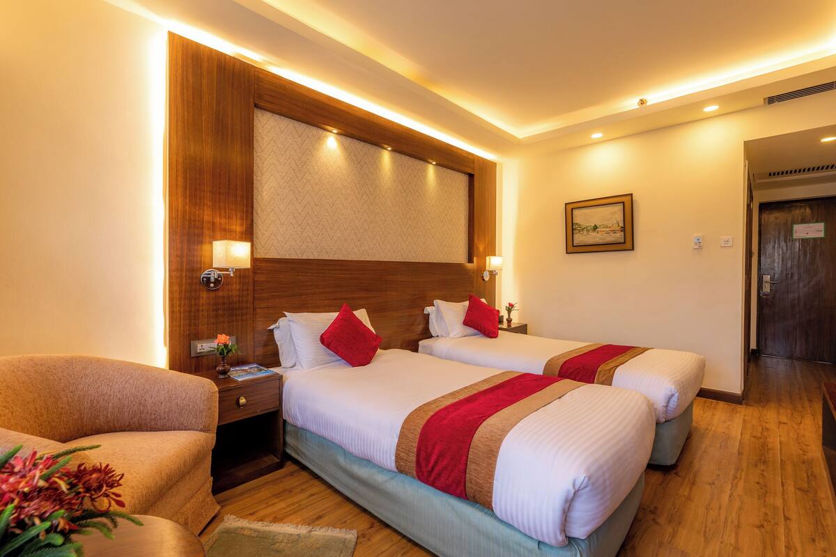 A hotel room with two beds, wooden headboard, bedside lamps, a nightstand, a chair, and a framed picture on the wall.
